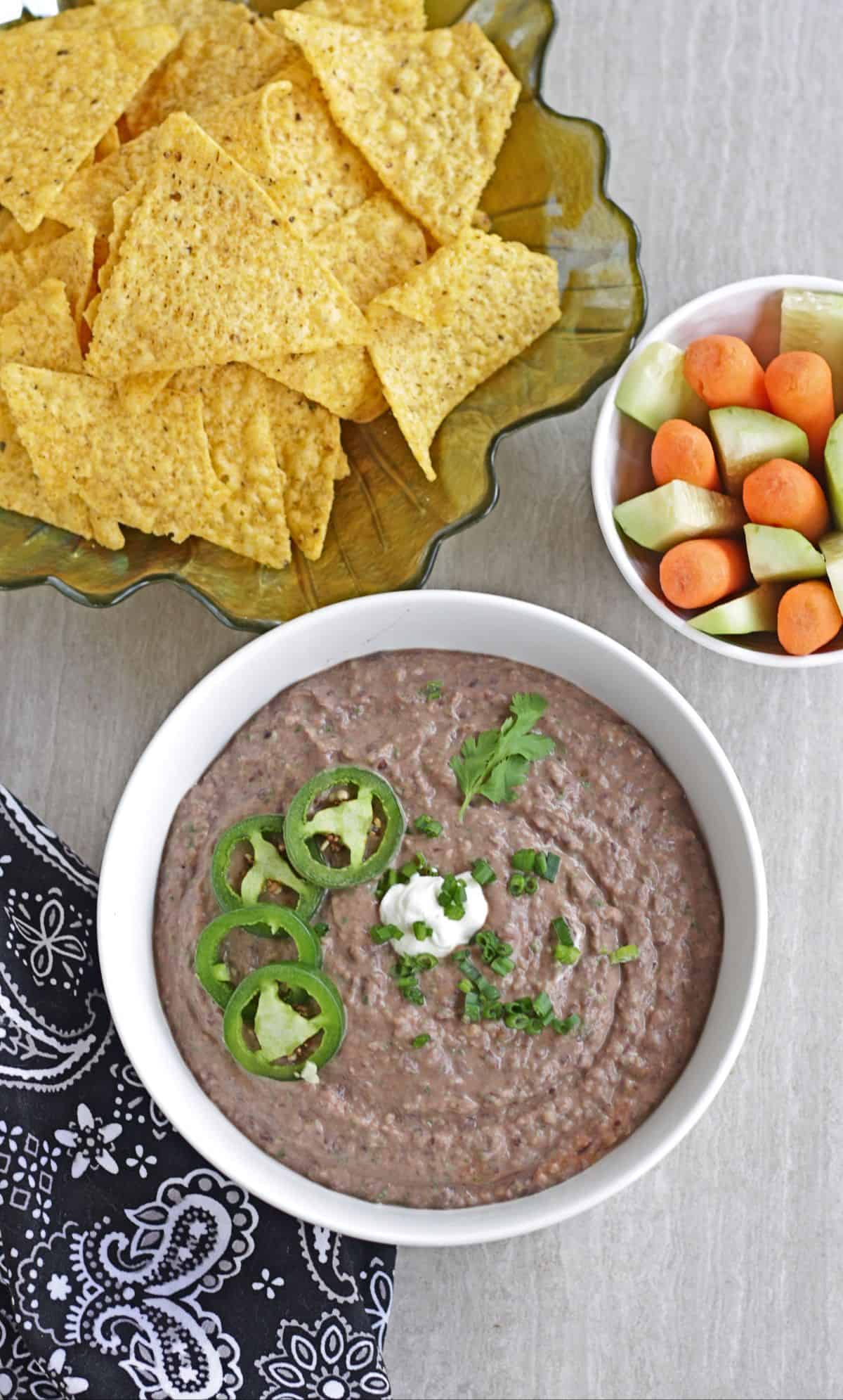 bean dip in a bowl with jalapeno garnish and chips and vegetables on the side