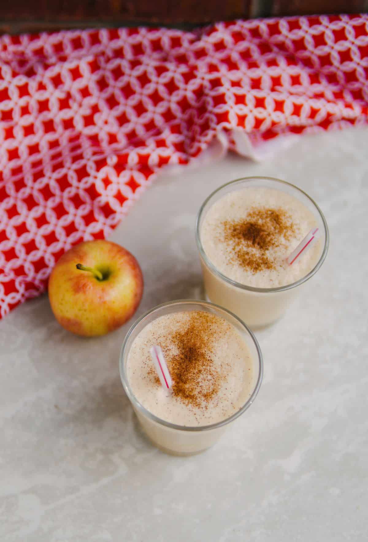 Two glasses of apple milkshake with a apple on the side.