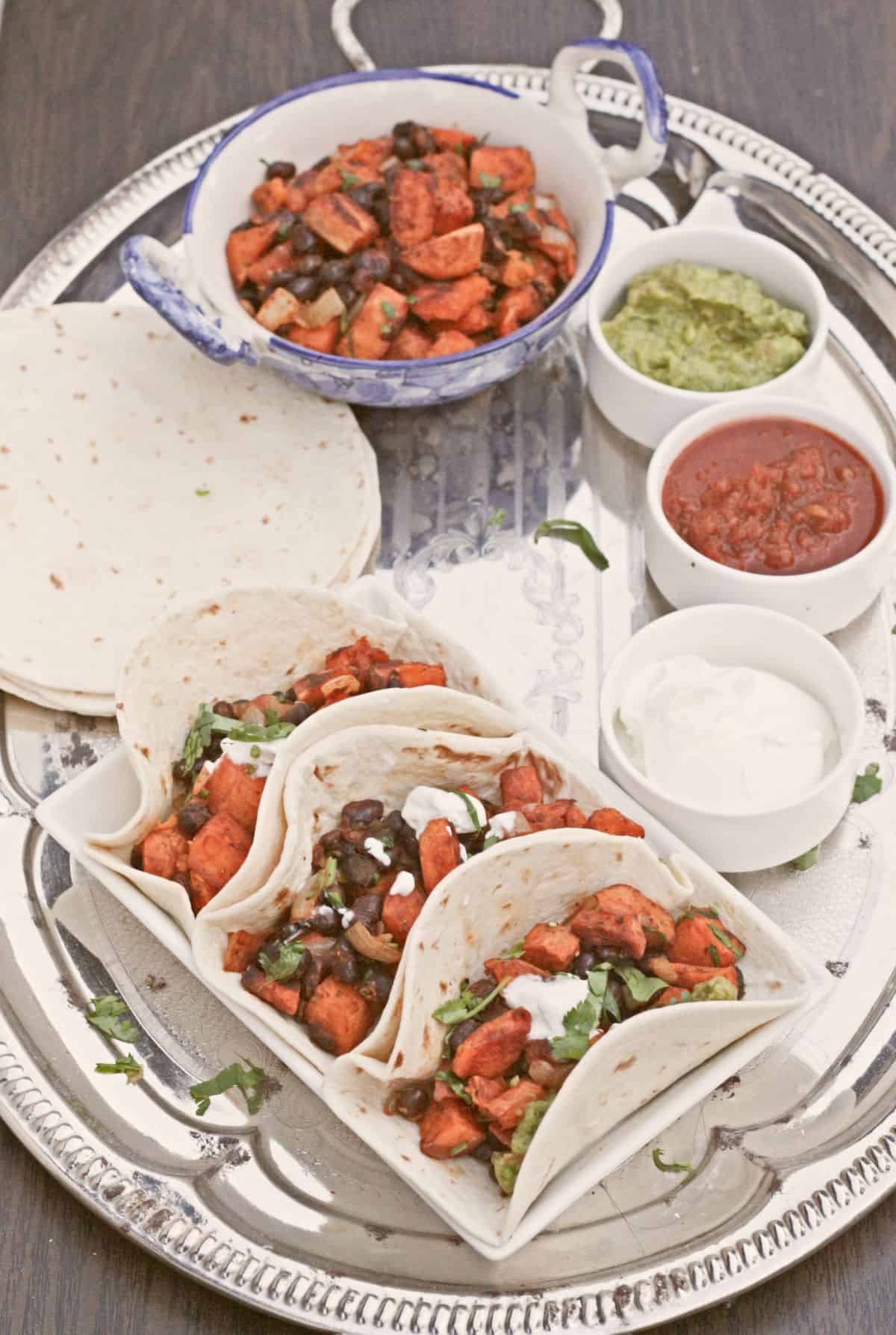 Soft tacos filled with black beans and sweet potatoes in a tray with sides.