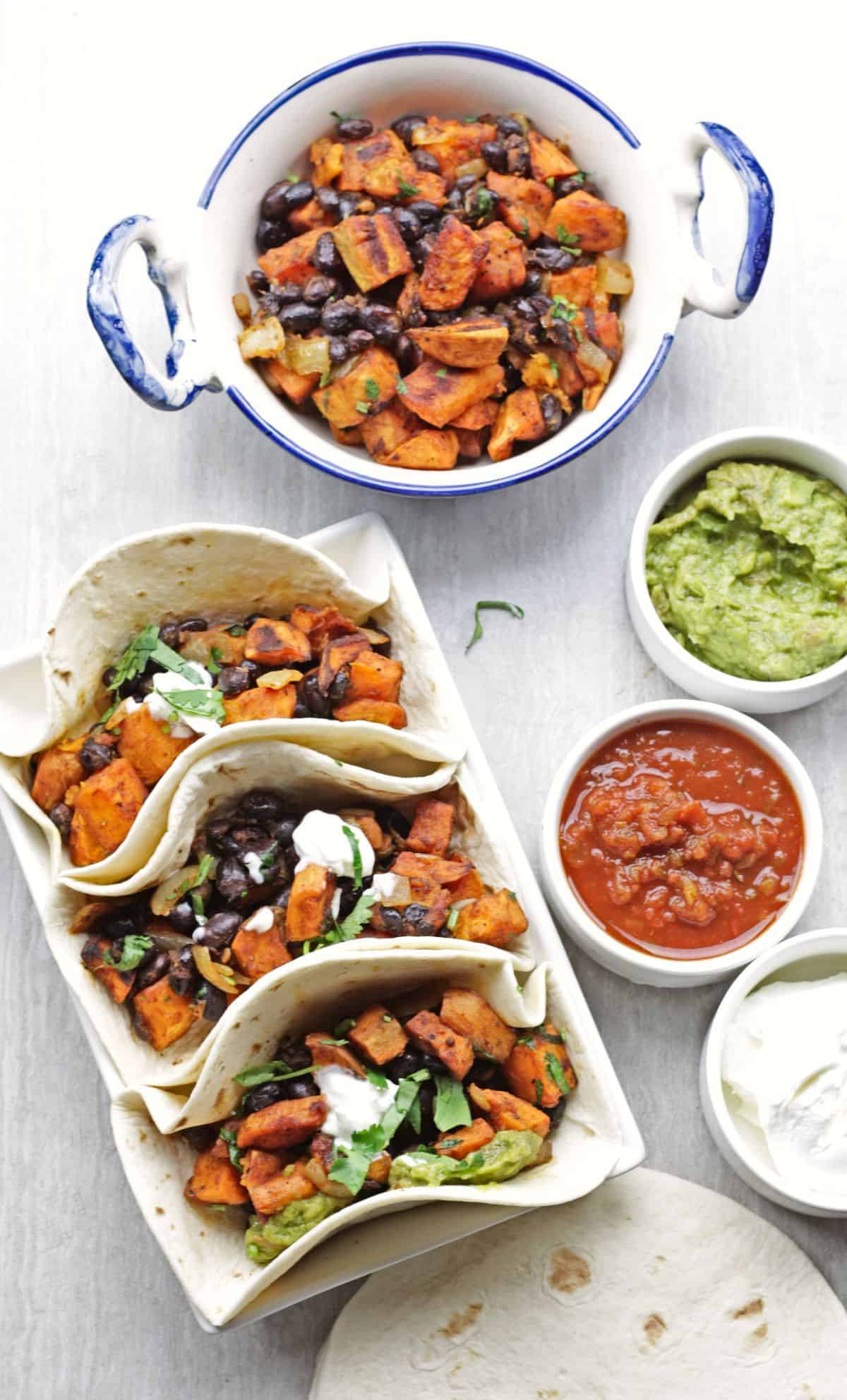 three soft tacos with black beans and sweet potato and salsa, sour cream and guacamole on the side.